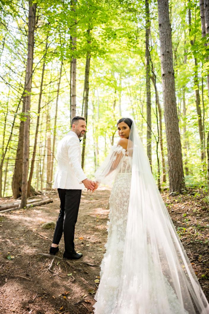 A noiva e o noivo dão as mãos e olham por cima dos ombros em seus trajes de casamento enquanto estão na floresta.